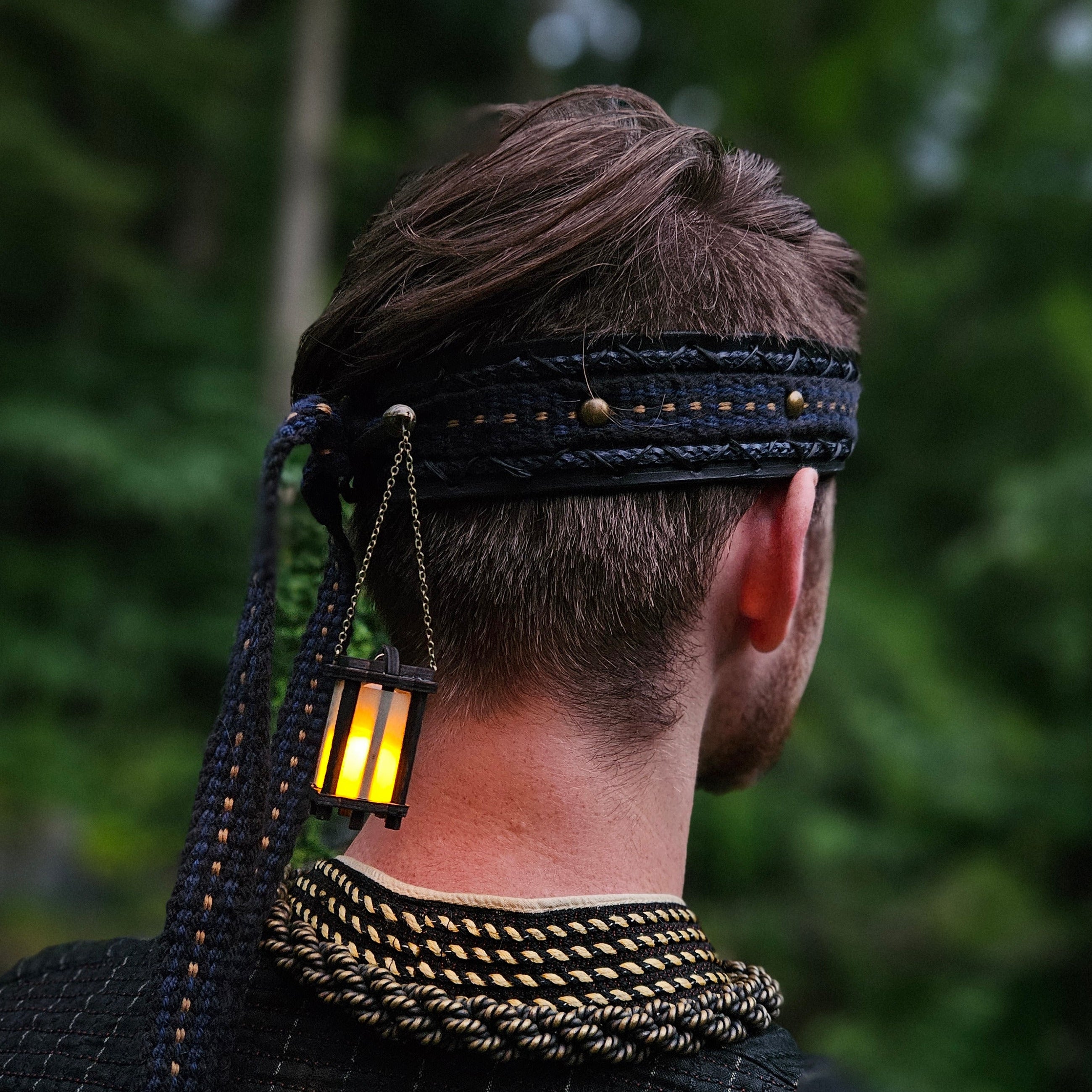 Person wearing a dark outfit with gold accents, including a headband with a lit glowing lantern dangling from it.