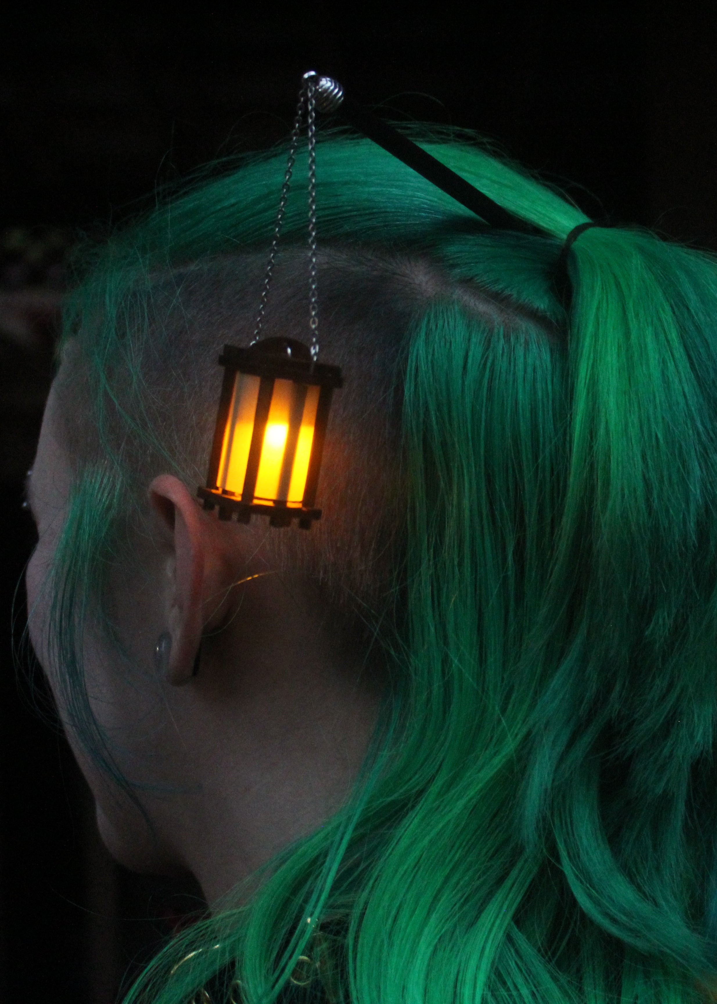 Person with green hair wearing a small glowing lantern on their head against a dark background